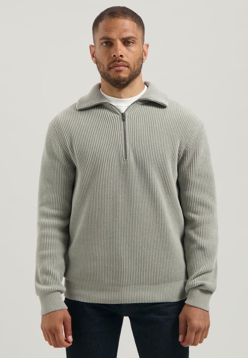 Man in een lichtgrijs geribbeld gebreid zip-up sweater met donkere broek, staande tegen een effen lichte achtergrond.