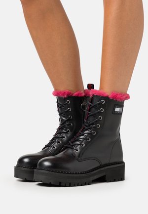 Bottes en cuir noir à lacets avec semelles épaisses et bordure en fourrure rose vif, portées sur des jambes nues, montrant une fermeture éclair sur le côté et un patch de la marque sur la cheville.