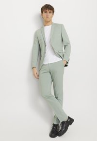 Lys grønn dress med slim fit, en-knaps jakke, matchende bukser og en hvit t-skjorte. Svart slitesterke sko fullfører antrekket.