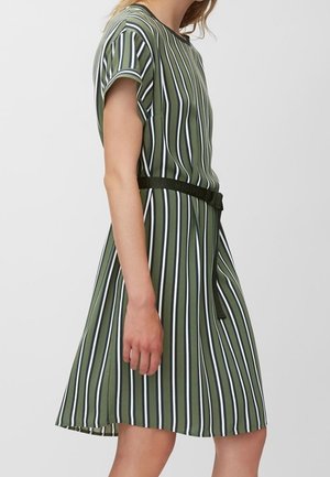 Kobieta ubrana w zieloną sukienkę do kolan z pionowymi białymi i czarnymi paskami oraz czarnym paskiem związanym w talii, pokazana z profilu.