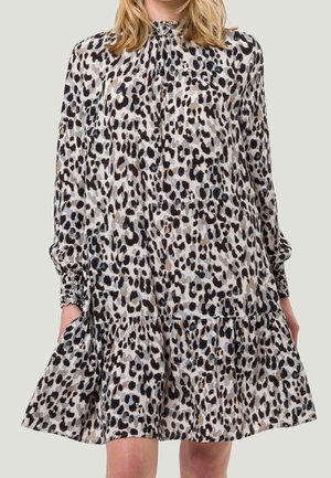 Kobieta w długiej sukience z długim rękawem, wysokim kołnierzem i luźnym zwierzęcym wzorem, z falbaną na dole, trzymająca boki spódnicy.