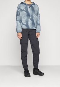 Leichte Jacke mit Kapuze in Blau- und Grautönen, kombiniert mit schwarzen, schmalen Hosen und schwarzen Schuhen. Der Fokus liegt auf der Funktionalität für den Outdoor-Bereich.