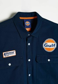 Granatowa koszula z długim rękawem z dwoma kieszeniami na piersiach, białymi guzikami oraz naszytymi emblematami "Gulf" i "Diverse ExtremeTeam."