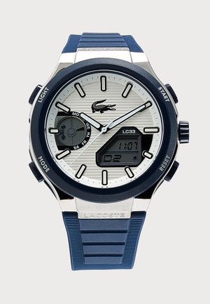 Montre-bracelet ronde argentée et bleue Lacoste avec aiguilles analogiques, indicateur du jour, écran digital et bracelet en caoutchouc bleu.