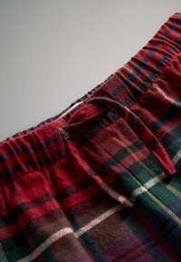 Tessuto a quadri rosso e verde con una texture morbida, caratterizzato da una vita elastica e da un dettaglio con fiocco sul davanti. Vista ravvicinata dell'area della vita.