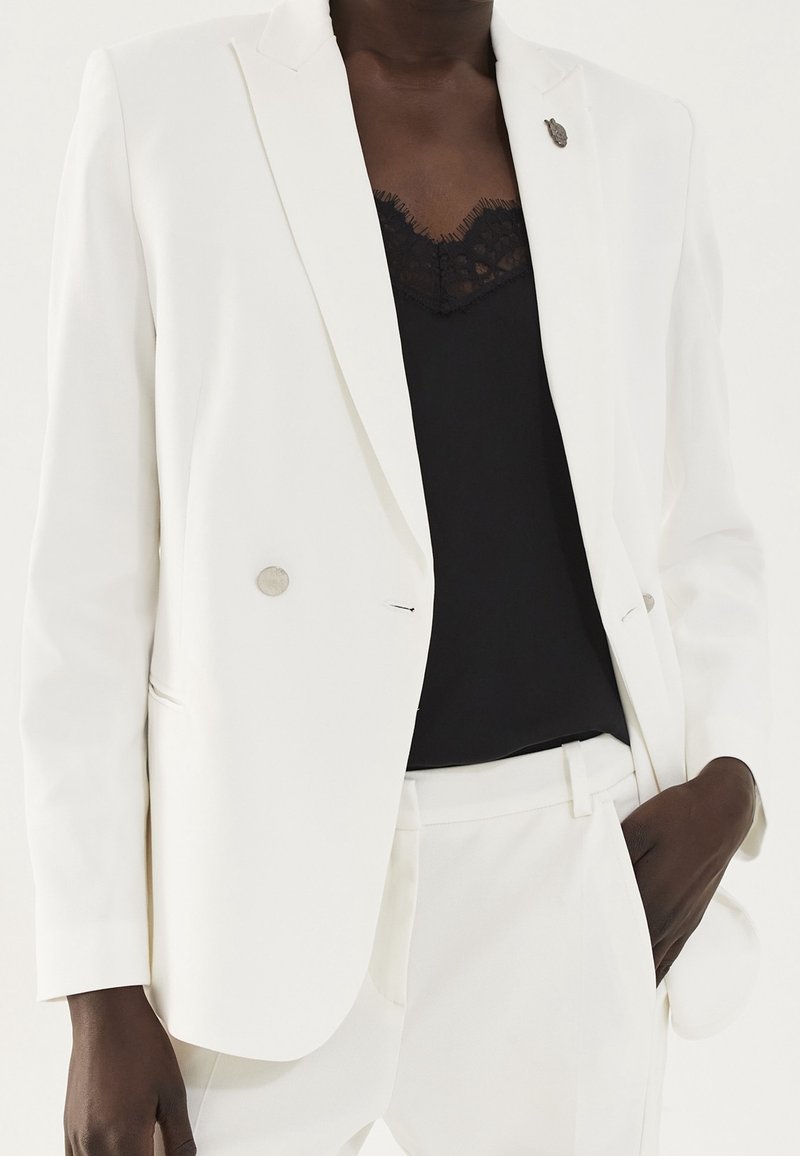 Femme portant un tailleur pantalon blanc à double boutonnage avec un caraco noir orné de dentelle, une main dans la poche, sur un fond uni.