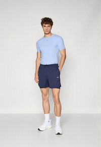 Hellblaues, tailliertes Kurzarmoberteil kombiniert mit marineblauen Shorts mit Kordelzug. Das Model trägt weiße Sneaker und Knöchelsocken und präsentiert so einen lässigen, sportlichen Look.