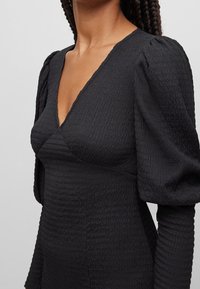 Robe noire texturée avec un décolleté en V et des manches bouffantes. Le tissu présente un motif légèrement froissé, accentuant sa silhouette structurée.