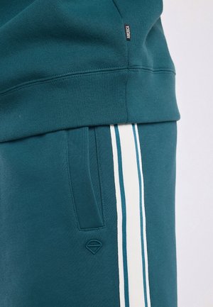Petrolkleurige joggingbroek met witte en petrolkleurige verticale zijstrepen, zijzak en een klein gestikt logo onder de zak; zichtbare zoom van bijpassende petrolkleurige trui.