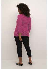 Maglione rosa lavorato a maglia con un motivo a onda testurizzato, maniche a tre quarti e un retro leggermente più lungo. Indossato con pantaloni neri a vita alta e scarpe nere.