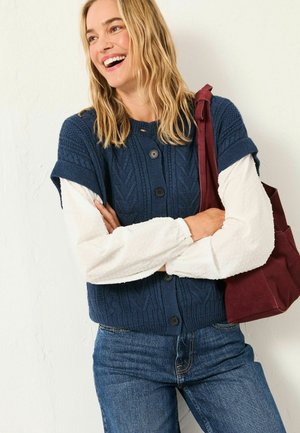 Navy knitted cardigan with textured pattern, short sleeves, and black buttons; worn over a white dotted blouse. Burgundy tote bag.