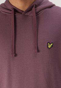 Sudadera morada con cordón y un pequeño logo de un pájaro bordado en amarillo en el pecho. El material parece suave y ligeramente texturizado.