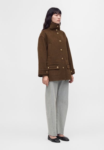 Femme portant un manteau marron boutonné avec col montant, jean ample gris clair et chaussures plates noires, debout devant un fond uni.