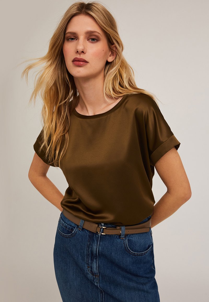 Mörk olivgrön satinkr tops med korta rullade ärmar, ihopparad med en jeanstygs kjol och ett brunt bälte, som visar en slät, glänsande textur.