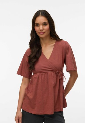 Naine pikkade tumedate juustega, seljas roostekarva lühikeste varrukatega mähistopp ja mustad püksid, seisab ühe käega selja taga.