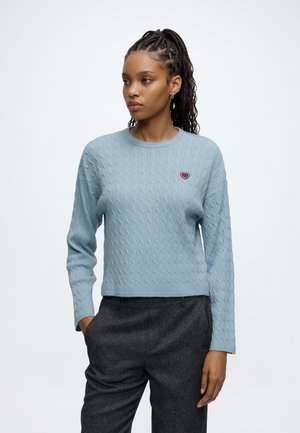 Femme aux cheveux tressés portant un pull en maille torsadée bleu clair avec un petit emblème en forme de cœur et un pantalon gris foncé, mains dans les poches, regardant vers la gauche.