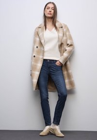 Beige kostkovaný vlněný kabát s černými knoflíky, nošený přes krémový svetr s V výstřihem, v kombinaci s tmavě modrými džíny a béžovými teniskami.