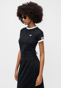 Μαύρο αθλητικό τοπ με λευκό γιακά και μανσέτες, με λεπτομέρεια πλαϊνής ρύθμισης με κορδόνι, και μικρό κεντημένο λογότυπο στο στήθος.