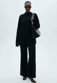 Crni oversize pulover s visokim ovratnikom, uparen s crnim hlačama s izrezom. Dodaci uključuju crne sunčane naočale i strukturiranu crnu torbu.