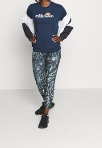 Vrouw die een marineblauwe ellesse-trui, een wit-zwarte jas, zwart met patroon bedrukte legging en zwarte sneakers draagt, staand met handen in haar zij.