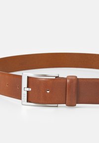 Bruine leren riem met een gladde textuur, voorzien van een metalen zilveren gesp. Verstelbaar met gaatjes voor de juiste pasvorm. Merkl logo gegraveerd aan de gespzijde.