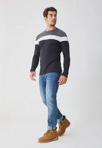 Zwart shirt met lange mouwen met grijze en witte strepen over de borst, gecombineerd met een blauwe jeans en bruine werkschoenen. Glad stoftextuur.