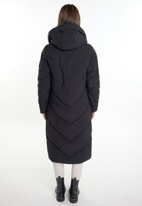Schwarzer langer Daunenmantel mit einem Chevron-Steppdesign, Kapuze und Reißverschlusstaschen, kombiniert mit hellen Hosen und schwarzen Stiefeletten.
