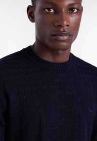 Mornarska pletena jopica z teksturo, ki ima okrogel izrez in subtilen modri logotip. Tkanina ima geometrijski vzorec in mehko površino.