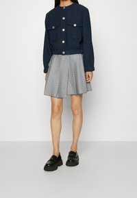 Giacca cropped blu navy con due tasche frontali e bottoni dorati, abbinata a una gonna svasata a righe bianche e nere, e mocassini neri con zeppa.