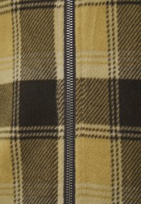 Fleecepiké med dragkedja som har ett gult och brunt rutmönster, med en metallisk dragkedja fram för stängning.