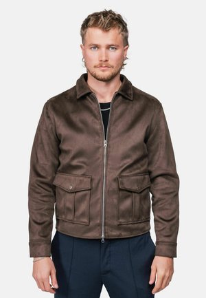 Homme aux cheveux courts et bouclés avec une barbe, portant une veste marron zippée avec poches à rabat et un pantalon bleu marine, debout devant un fond uni.