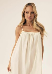 Helles beige ärmelloses Kleid mit gerafftem Rundhalsausschnitt, fließender Silhouette und weicher Textur. Einfaches Design mit dünnen Trägern.