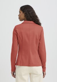Veste à col en tissu corail, coupe ajustée, manches longues, détails de coutures à l'arrière, associée à un pantalon de couleur claire.