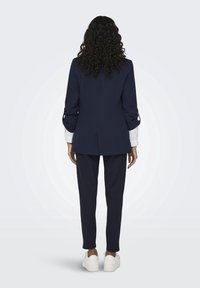 Blazer bleu marine au design structuré, manches retroussées et coupe ajustée, associé à un pantalon assorti et des baskets blanches. Vue de dos.