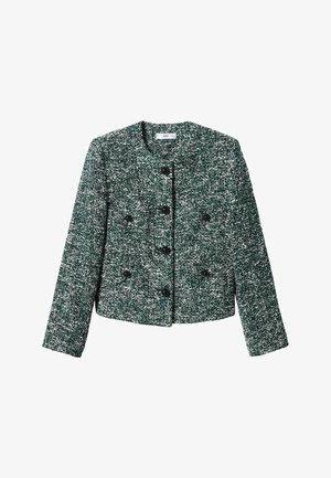 Donna che indossa una giacca verde texturizzata, una minigonna nera e stivali in pelle neri al ginocchio, in piedi su un pavimento neutro con sfondo bianco.