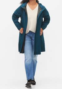 Tyrkosa regnjacka med huva, dragkedja och snörning, med sidofickor och en avslappnad passform, bärs över en ljusgrå tröja och blå jeans.