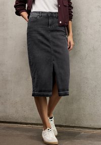 Mikati musta denim midi, jolla on etu halkio, klassinen viisi-taskumalli ja raaka helma. Yhdistetty valkoisten tennareiden kanssa betonitaustalla.