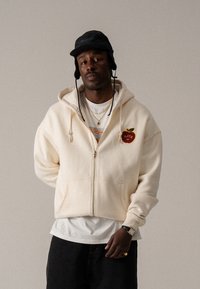 Beige φούτερ με φερμουάρ και τσέπη, διακοσμημένο με κόκκινο κεντημένο λογότυπο μήλου. Φοριέται πάνω από λευκό πουκάμισο και συνδυάζεται με σκούρα τζιν. Προστίθεται μαύρο καπέλο.