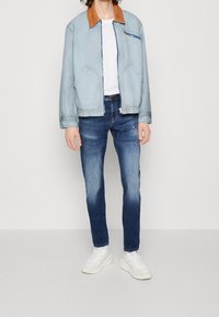 Chaqueta azul claro con un collar marrón, con un bolsillo con cremallera y bolsillos laterales, combinada con jeans azul oscuro y zapatillas blancas.