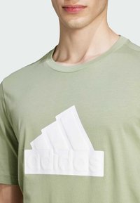 Camiseta de algodón verde claro con un gran logotipo de Adidas en relieve de color blanco en forma de tres franjas diagonales en la parte delantera.