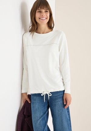 Top bianco a maniche lunghe con scollatura rotonda e orlo con cordino. Un testo stampato discreto adorna la parte frontale. Abbinato a jeans blu.