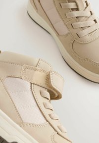 Baskets montantes beige avec une finition mate lisse, présentant un panneau latéral en maille texturée, un devant lacé et une sangle Velcro.