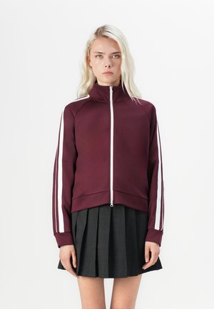 Burgundy zip-up jacket with high collar and white side stripes. Textured fabric, cropped design, paired with a grey pleated skirt.