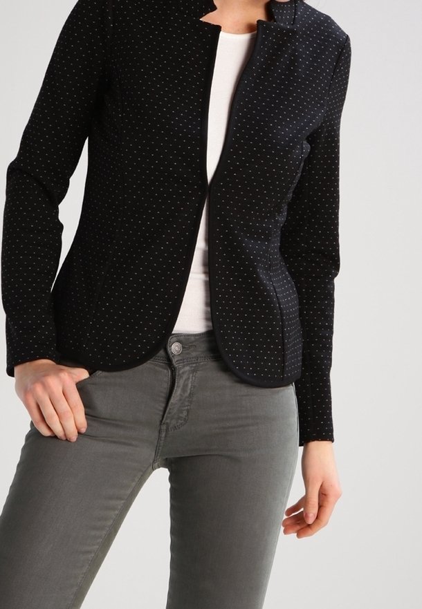 Černý blazer s bílými puntíky, strukturovaný design, klopový límec, slim fit. Kombinováno s bílým topem a šedými džíny.