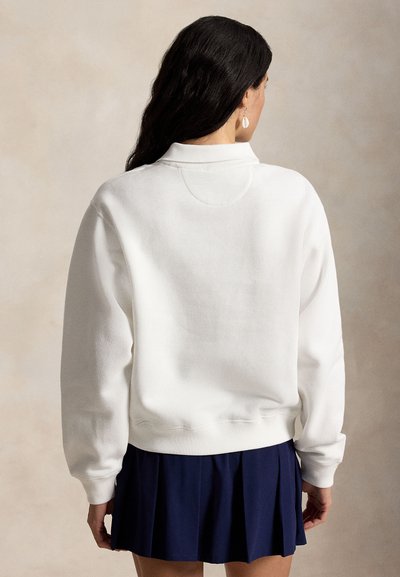 Femme aux longs cheveux foncés portant un sweat-shirt blanc et une jupe plissée bleu marine, vue de dos sur un fond neutre.