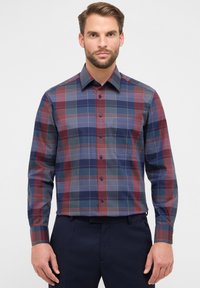 Homme portant une chemise à manches longues avec un motif à carreaux bleu, rouge et gris, rentrée dans un pantalon noir marine, se tenant devant un fond blanc.