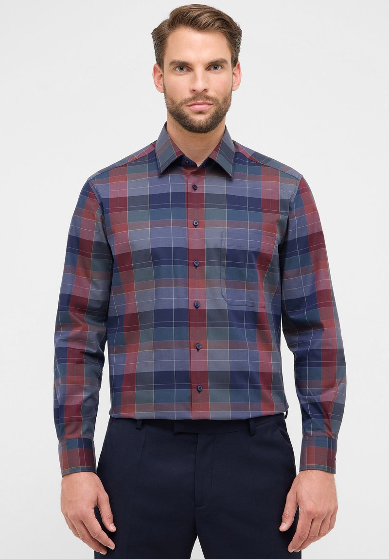 Homme portant une chemise à manches longues avec un motif à carreaux bleu, rouge et gris, rentrée dans un pantalon noir marine, se tenant devant un fond blanc.