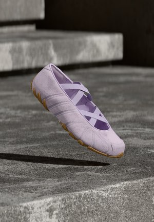 Lila instapschoenen met kruisvormige elastische bandjes en een gestructureerde zool, zwevend boven betonnen traptreden in een buitenomgeving.