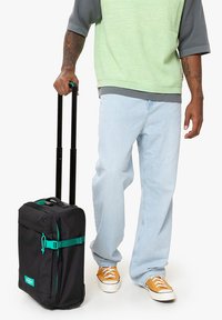 Černý kufr na kolečkách s tyrkysovými akcenty, obdélníkového tvaru, pevnou rukojetí a nastavitelnými teleskopickými kolečky, umístěný vedle světle modrých džínů a oranžových tenisek.