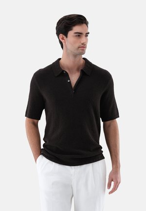 Mann mit dunklem Haar, der ein schwarzes kurzärmeliges, strukturiertes Poloshirt und weiße Hose trägt, steht mit den Händen in den Taschen und schaut zur Seite.